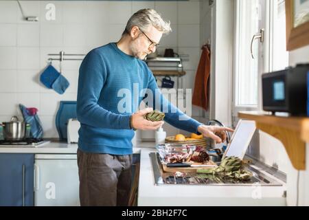 Mature man preparing salad in his kitchen using digital tablet Stock Photo
