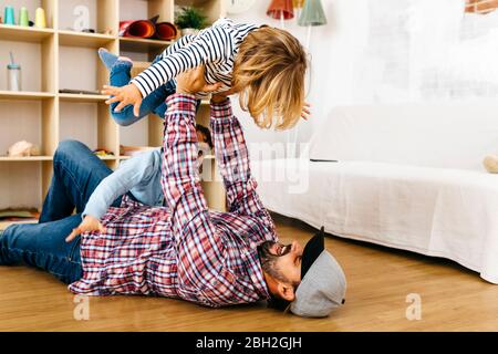 Father lying on floor, holding daughter aloft, pretending to fly Stock Photo