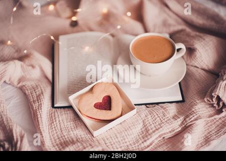 Fresh cup of coffee with heart shape cake on open book in bed over lights closeup. Good morning. Breakfast. Valentines Day.