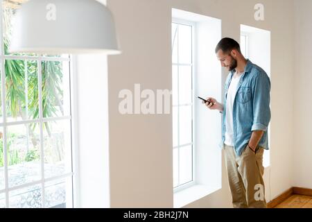 Young man at home checking his smartphone and standing in front of his windows in modern living room Stock Photo