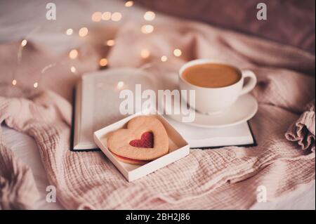 Fresh coffee in white cup staying on p[en book with heart shape cake over glowing lights closeup. Good morning. Valentines Day. Stock Photo