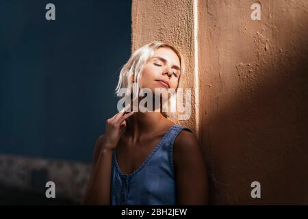 Portrait of young blond woman leaning against wall enjoying sunlight Stock Photo