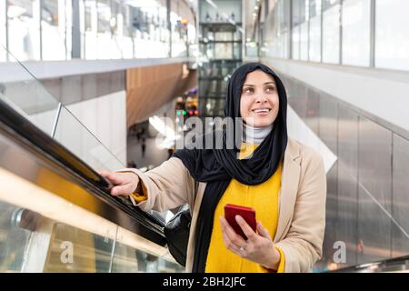 Portrait of a smiling young woman wearing hijab standing on escalator Stock Photo