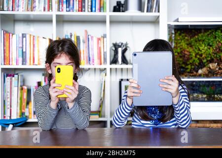 Two sisters sitting side by side at table in the living room using electronic devices Stock Photo