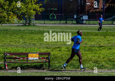 London, UK. 23rd Apr, 2020. Running past a closed bench - Clapham Common is reasonably busy as the sun is out and it is warmer. The 'lockdown' continues for the Coronavirus (Covid 19) outbreak in London. Credit: Guy Bell/Alamy Live News Stock Photo