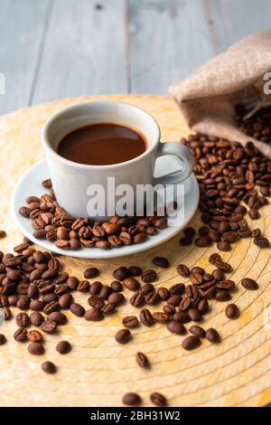 White cup and saucer with coffee and surrounded by roasted coffee beans on a wooden table and a bag Stock Photo