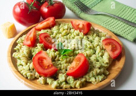 Basil pesto fusilli pasta with Parmesan cheese and sliced tomatoes in a wooden plate and a green napkin with a fork in the background