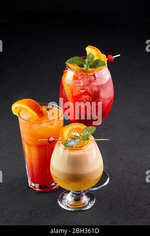 Three alcoholic cocktails, red, orange and yellow colors on black backdrop. Variety of alcoholic drinks decorated with orange slices Stock Photo