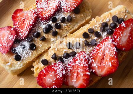 Peanut butter spread toasts with chocolate chips, strawberries, coconut flakes and homemade bread on a wooden plate Stock Photo