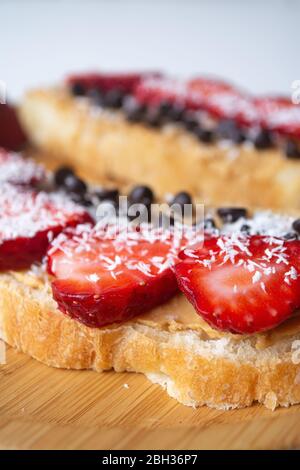 Peanut butter spread toasts with chocolate chips, strawberries, coconut flakes and homemade bread on a wooden plate Stock Photo