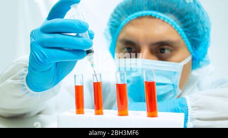 Close up view of  latin  doctor in bioprotective suit, face mask and blue gloves taking a sample of red liquid from a glass test tube with a syringe o Stock Photo