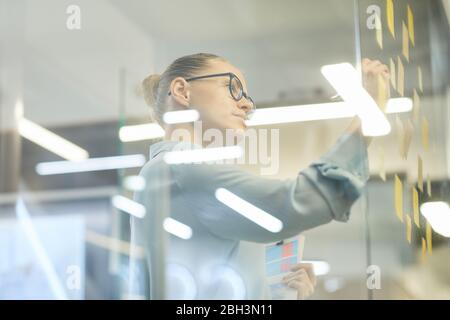 Young businesswoman in eyeglasses making notes on adhesive notes on the glass wall at office Stock Photo
