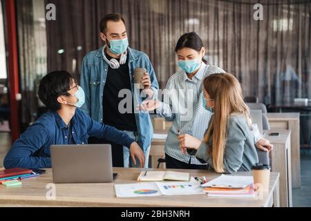 Image of multinational joyful students in protective mask talking while studying with laptop at classroom Stock Photo
