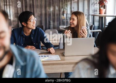 Photo of multinational joyful students talking and using smartphone while studying at classroom Stock Photo