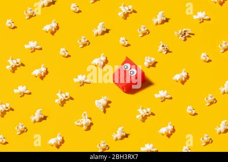 Empty popcorn box carton with googly eyes surrounded by popped popcorn on a yellow background Stock Photo