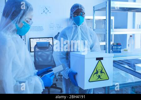 African chemist in protective workwear holding box with chemical samples with woman who using digital tablet while they working in the lab Stock Photo
