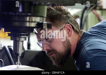Drill press operator examines drill bit in factory using safety glassesproduction Stock Photo