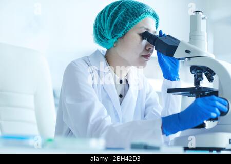 Asian young woman in white coat sitting at the table and examining analysis with microscope at office Stock Photo