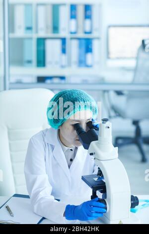 Female doctor in white coat sitting at the table and looking through the microscope and examining the samples at hospital Stock Photo