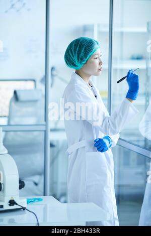Female scientist in white coat writing plans on glass wall while working at office Stock Photo