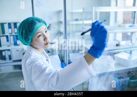 Asian female doctor in white coat and protective gloves making notes on glass wall at office Stock Photo