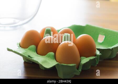 Six eggs sitting in a green cardboard tray open on a wooden counter top. A mixing bowl can be seen in the background Stock Photo
