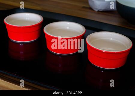 Three red ramekins sitting on a black tray. The black tray is on top of a wooden counter top with general kitchen items in the back of the photo Stock Photo