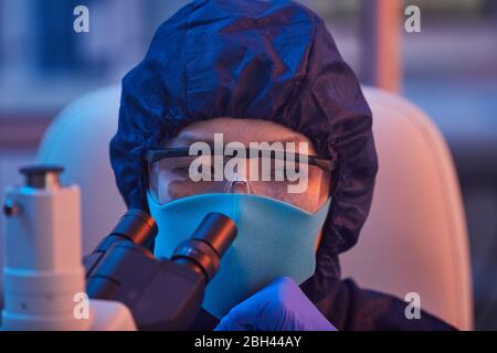 Close-up of female scientist in protective mask and glasses looking through the microscope while working at the lab Stock Photo