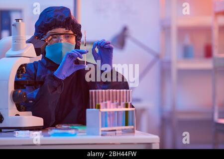 Female scientist in protective mask sitting at the table with test tubes and microscope and she using syringe at the lab Stock Photo