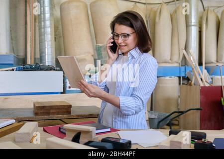 Female carpenter designer working in carpentry workshop, choosing materials, talking on phone. Industrial business, woodwork Stock Photo