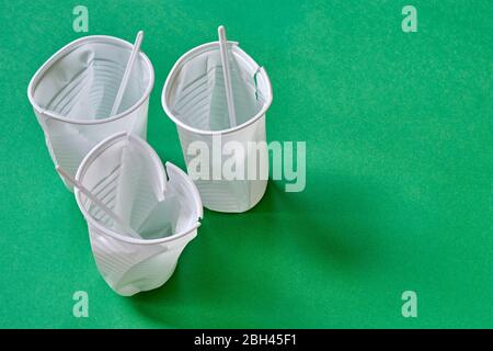 Three standing smashed white plastic coffee cups with stirrers on a green background with copy space. Zero waste, plastic free, stop pollution. Stock Photo
