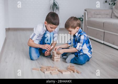 Two brothers boys build a tower of wooden blocks of the house on the floor. Table game. The concept of independent family quarantine, infection, coron Stock Photo
