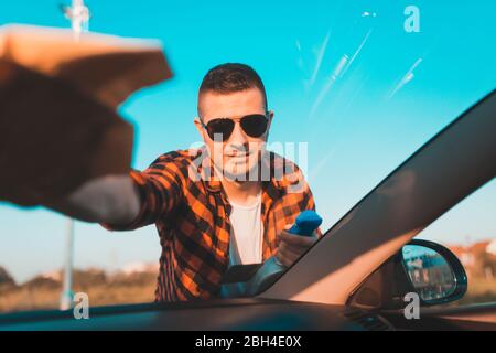 Young man carefully cleaning a windshield on his car with microfiber cloth and spray in a bottle. Stock Photo