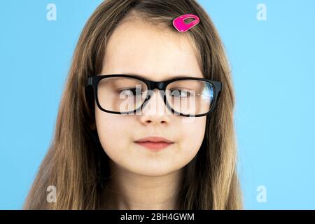 Close up portrait of a cross eyed child school girl wearing looking glasses isolated on blue background. Stock Photo