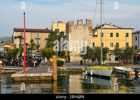 View of the old town of Bardolino on the shore of Lake Garda with the medieval tower and people in outdoor café in a sunny winter day, Verona, Italy Stock Photo