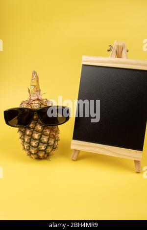 Pineapple wearing sunglasses with blank black board on a yellow background Stock Photo