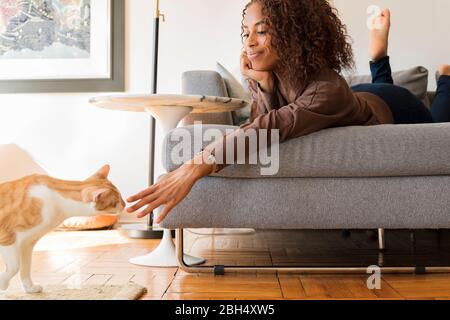 Smiling woman on sofa reaching out to cat Stock Photo