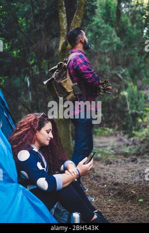 Woman sitting in tent using smart phone by man holding sticks Stock Photo