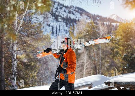 Man in orange jacket holding ski and looking at snow mountain at sunny day Stock Photo