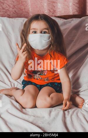 Sick little girl in mask shows on her fingers a sign staying at home in quarantine 2021 Stock Photo