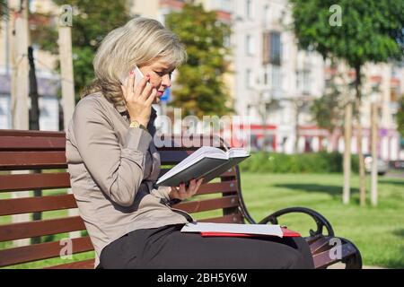 Business confident mature woman talking on cell phone, successful female in city sitting on bench, looking at business notebook Stock Photo