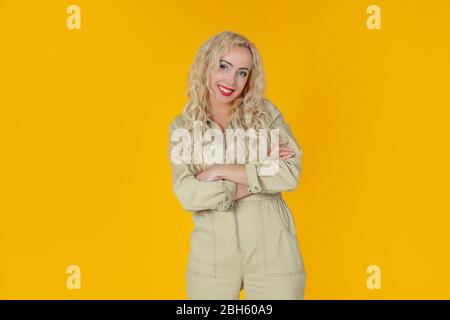 Portrait of a joyful and cheerful cute European curly-haired blonde woman who jokes cheerfully, entertains the conversation, smiles and laughs at the camera over the yellow wall. What a wonderful day Stock Photo