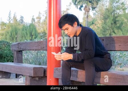 A boy in black clothes sitting in Thai local train station and watching his phone. Stock Photo
