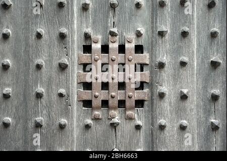 Viewing window slot with protecting rusty metal strips crisscrossing on ancient wooden door panel studded with rows of wooden beads. Stock Photo