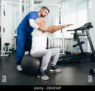 senior patient having physical therapy appointment with his doctor . Man doing post rehabilitation exercise while sitting on a fitness ball. Stock Photo