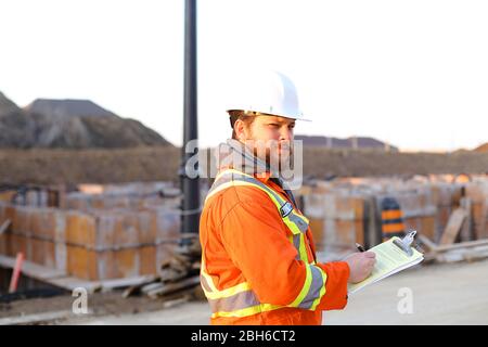 Caucasian inspector in orange workwear with blueprints on construction site. Stock Photo