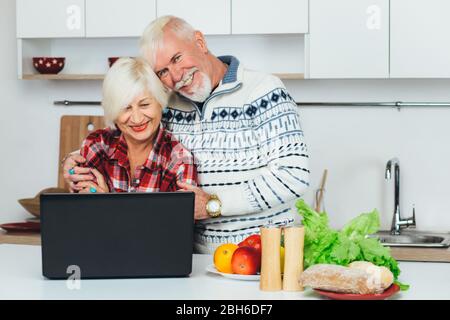 Elderly man and woman are looking for a recipe in a laptop, preparing healthy food at home, in the kitchen Stock Photo