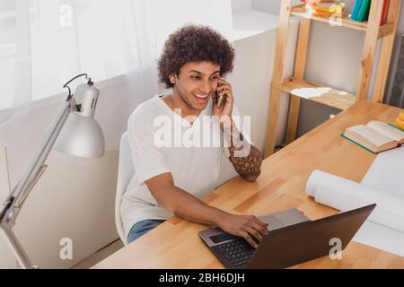 attractive and Curly mulatto man, surf the Internet, on his laptop and emotionally talking on the mobile phone while sitting at his working place at o Stock Photo