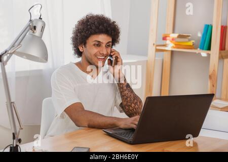 Pleasant conversation. Positive and Curly mulatto man, surf the Internet, on his laptop and Emotionally talking on the mobile phone while sitting at h Stock Photo
