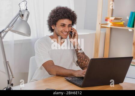 Have a pleasant chat. Positive and Curly mulatto man, surf the Internet, on  his laptop and talking on the mobile phone while sitting at his working p Stock Photo